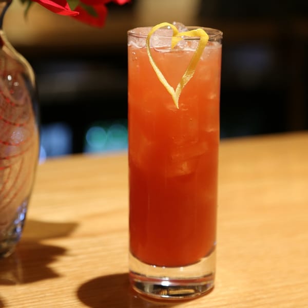 A tall glass of Sakanaya's red cocktail, garnished with a heart-shaped lemon peel, on a wooden bar.