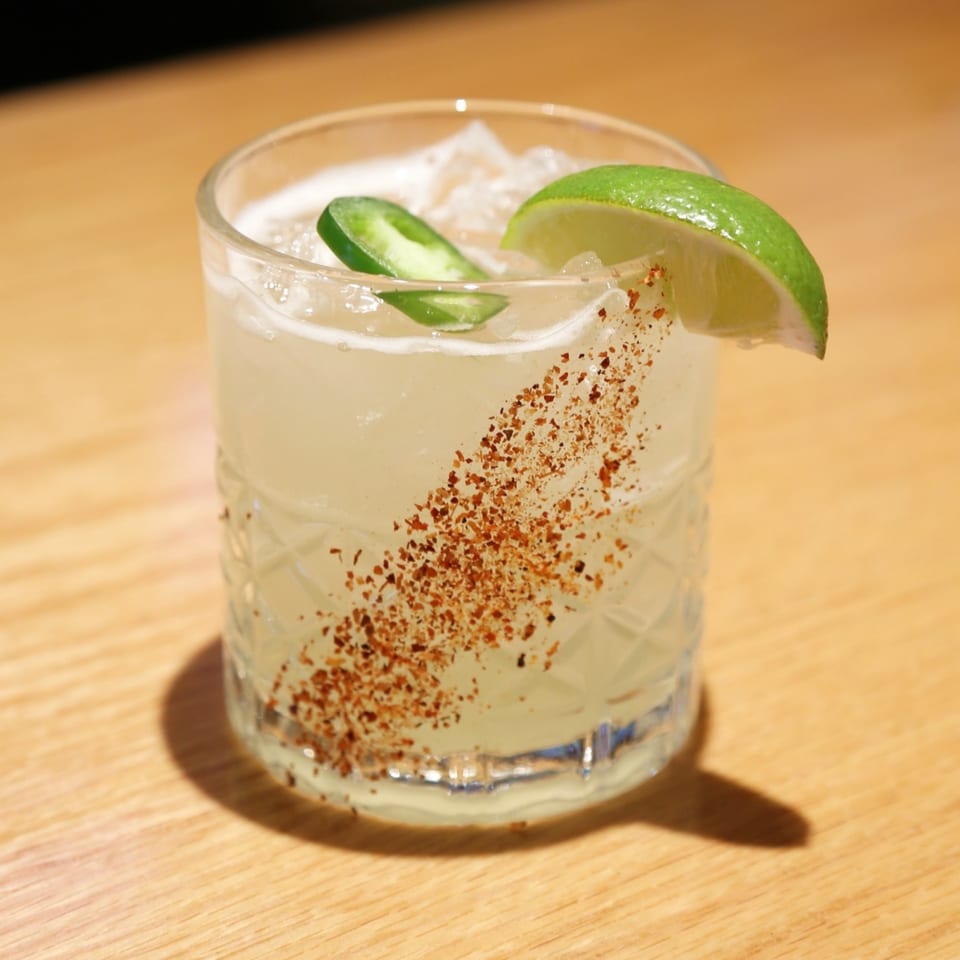 A Sakanaya cocktail with lime, jalapeño, and chili flakes in a textured glass on a wooden table.