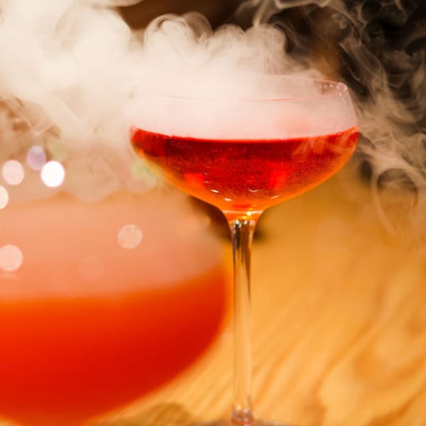 A red cocktail in a coupe glass with white smoke rising from it, on a wooden table.