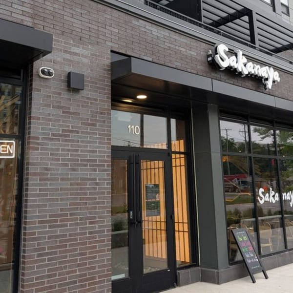 Exterior of Sakanaya, a Japanese restaurant with a brick facade, large windows, and a black awning.