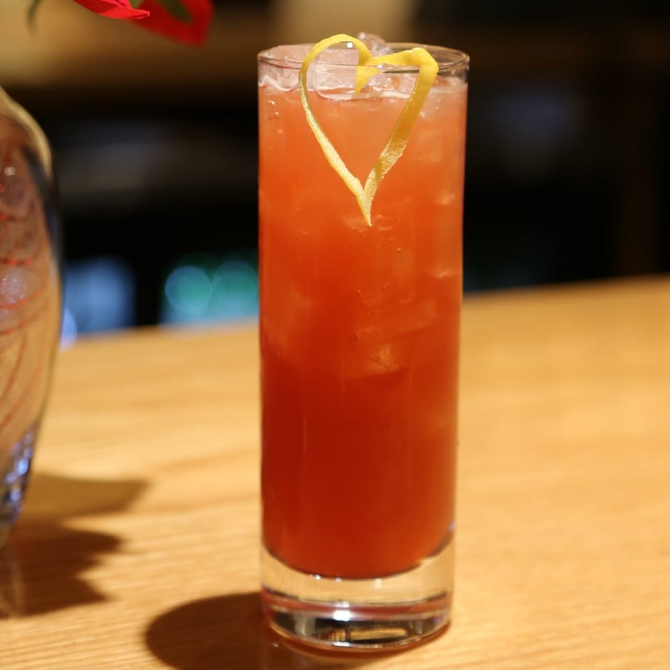 A tall glass of red cocktail with ice and a heart-shaped lemon peel garnish on a wooden table.