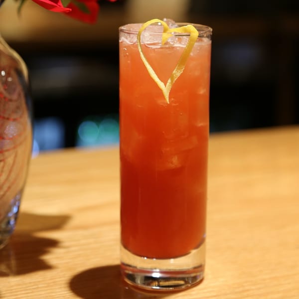 A tall glass of Sakanaya's red cocktail with ice and a heart-shaped lemon peel garnish.