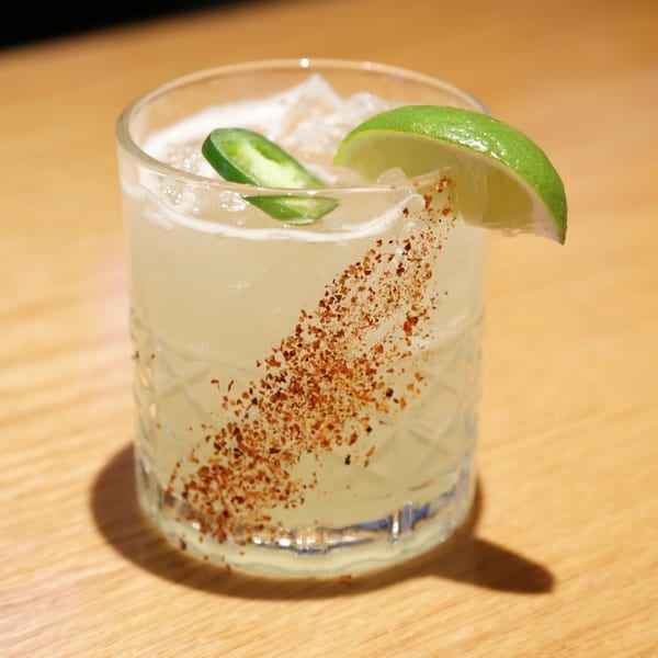 A Sakanaya cocktail with lime, jalapeño, and chili flakes in a textured glass on a wooden table.
