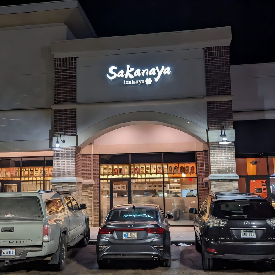 Exterior night view of Sakanaya Japanese restaurant with its glowing sign and parked cars.